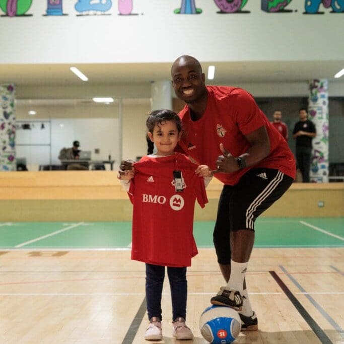 Soccer coach in red shirt and black pants stands next to young soccer player holding up a red jersey. Coach has one foot up on a soccer ball.