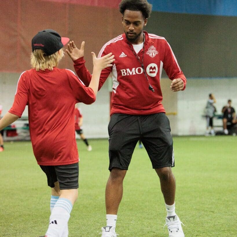 A soccer coach in a red Toronto FC jacket gives a high five to a young player wearing a red shirt, black shorts, and a black cap during an indoor training session on artificial turf.